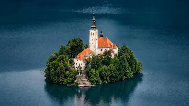 High angle aerial shot of a small green island with a red-roofed church centered in deep blue lake water.
