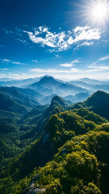 High altitude aerial view of lush green mountain peaks and valleys bathed in bright sunlight under a blue sky.