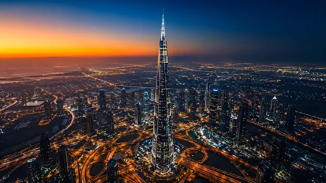 Aerial 3D render of an iconic supertall glass skyscraper towering over a sprawling city network of golden lit highways at dusk.
