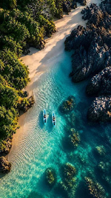 An aerial drone view of two blue kayaks paddling through clear turquoise water between a sandy beach and rocky cliffs.