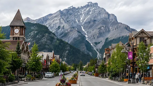 A bustling mountain town street lined with shops, cars, and trees, centered directly in front of a giant towering mountain peak.