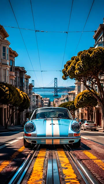 A light blue classic sports car with white racing stripes sits on a steep paved street with cable car tracks leading to a bridge in the distance.