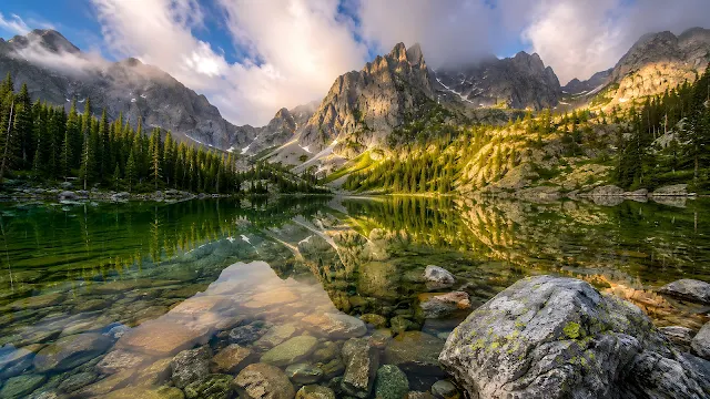 A glass-like clear alpine lake reflecting jagged snow-patched mountains under soft overcast clouds with submerged rocks in the foreground.