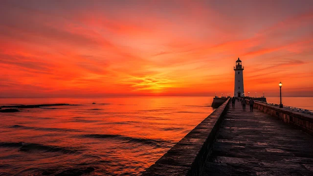 A white lighthouse stands at the end of a stone pier extending into a calm sea under a vibrant pink-orange sunset.