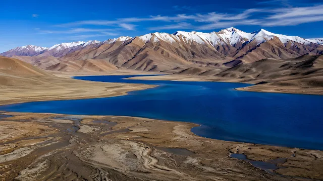 An elevated view of a deep blue high-altitude lake with dry, brown mudflats in the foreground and rugged mountains in the distance.