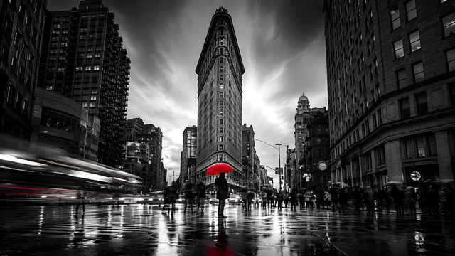 Monochrome view of the Flatiron Building in New York City with a person holding a red umbrella on wet pavement.