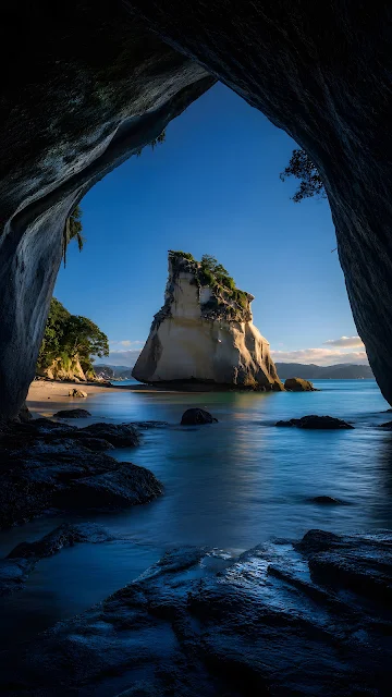 View looking out from inside a dark coastal rock cave frame toward a dramatic sea stack island in clear blue water.