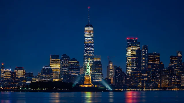 An illuminated harbour monument stands before a glowing skyline across calm blue water at night.