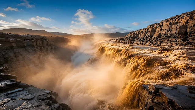Powerful muddy brown water cascades through a misty rocky canyon under warm daylight.