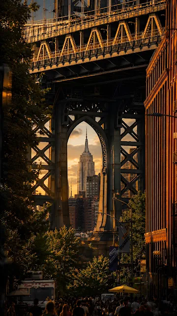 Looking through a massive steel bridge tower arch to reveal a distant art deco skyscraper framed perfectly at golden hour.