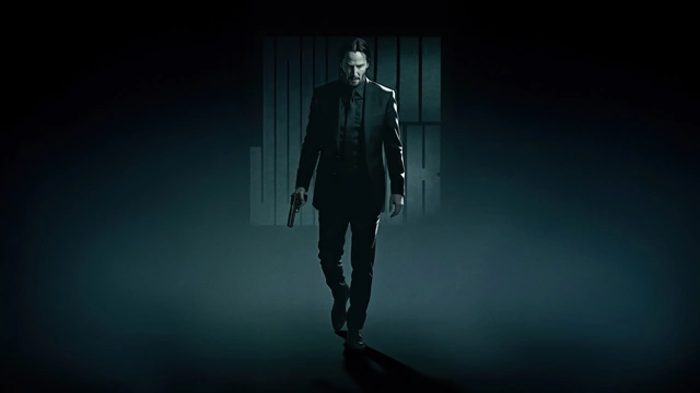 John Wick walking forward in a dark suit holding a handgun in his right hand.