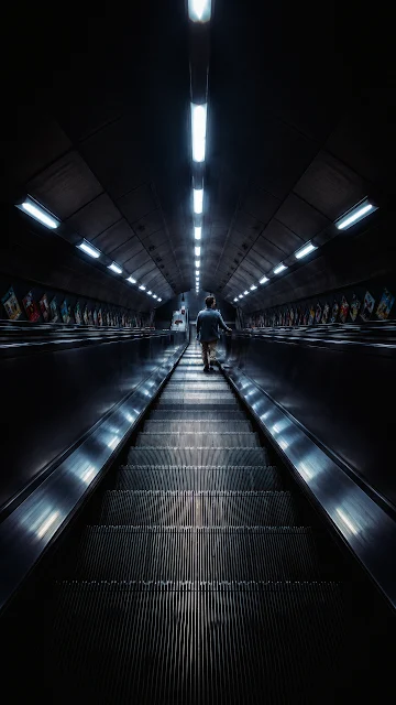 A lone commuter seen from behind standing on a long metallic escalator inside a dark subway tunnel.