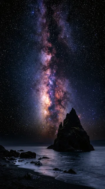 Vibrant glowing Milky Way galactic core arching over a dark monolithic coastal sea stack in pitch-black night.
