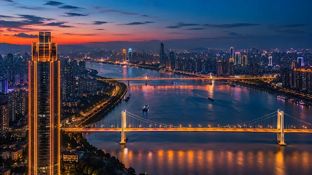 High-angle night view of a sprawling illuminated modern city along a wide river with glowing suspension bridges at sunset.