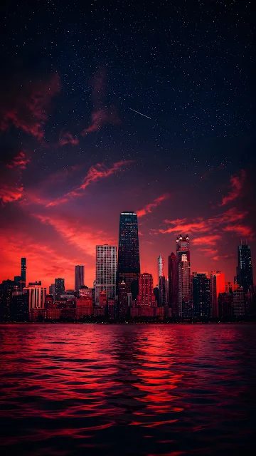 A panoramic city skyline silhouette under a dramatic vibrant crimson red sunset sky reflected on shimmering water in the foreground.