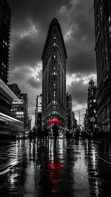 Monochrome street view of a triangular historic skyscraper on a rainy day with a person carrying a bright red umbrella.