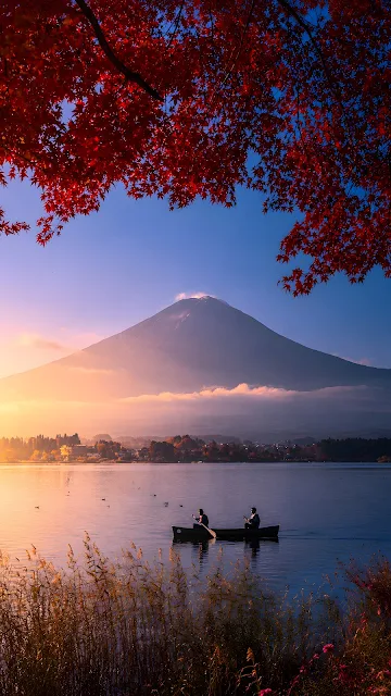 Vertical landscape photograph of Mount Fuji framed by fiery red maple leaves above a calm lake with two people in a small canoe at sunrise.