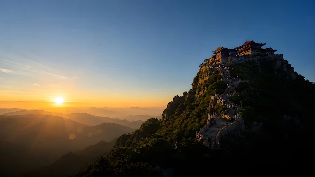 A stone path crowded with visitors leads to a mountain peak temple against a brilliant rising sun flare.