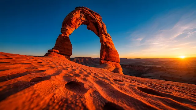 Massive natural red rock arch over rippled sand dunes illuminated by a low setting sun in a desert landscape.