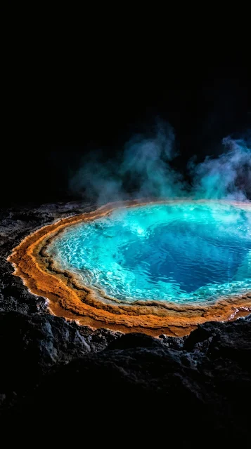 Vibrant glowing neon turquoise and orange geothermal mineral pool surrounded by pitch-black volcanic rock at night.