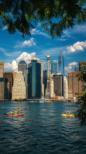 Kayakers paddle across blue water in front of the towering Lower Manhattan skyline under a partly cloudy sky, framed by tree leaves.