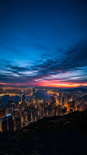 High angle panoramic view of a massive modern city harbor skyline lit up at dusk beneath a dramatic sunset sky.