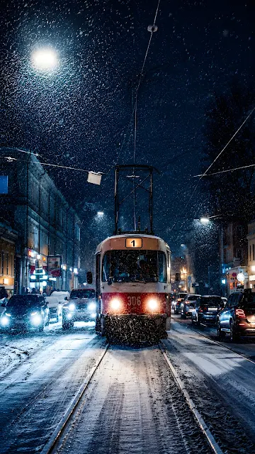 Low angle long exposure photograph of a vintage red and white streetcar moving down wet snow-covered tracks in a European city street at night during heavy snowfall.
