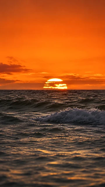 Close-up view of dark ocean waves with white sea spray beneath an intense bright orange sunset sky.