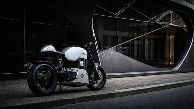 White Savic electric motorcycle parked on a concrete curb in front of an architectural modern building with large curved glass.