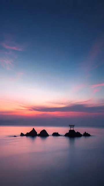 Small traditional torii gate perched on rocks in a smooth sea under a soft pink and blue sunset sky.