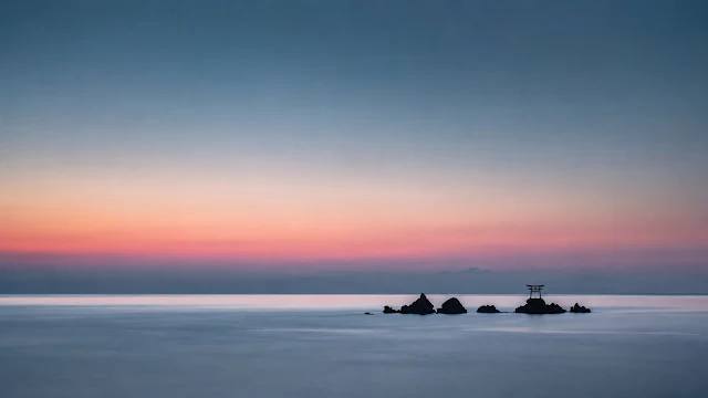 Small black torii gate perched on offshore rocks in calm blue ocean water beneath a pink sunset gradient sky.