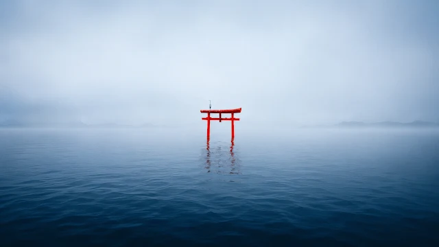 A vibrant vermilion torii gate standing in the center of still blue water surrounded by soft white morning fog.