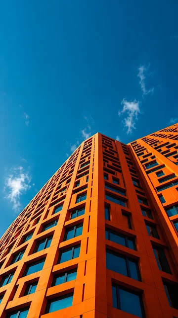 Low angle view looking up at a vibrant orange modern skyscraper against a clear blue sky with negative space.