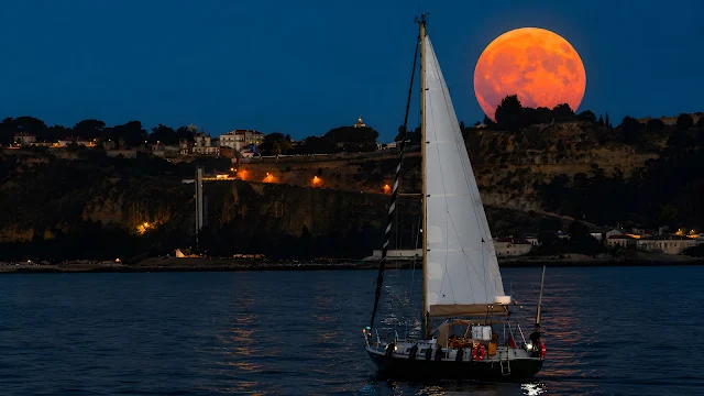 A white sailboat with raised sails navigating calm water at dusk with a massive reddish-orange full moon behind a coastal hill.