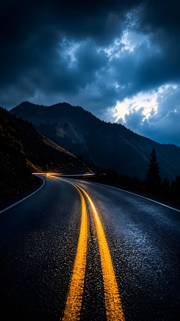 S-curve mountain road at twilight with double yellow lines in the foreground leading into dark mountains under storm clouds.